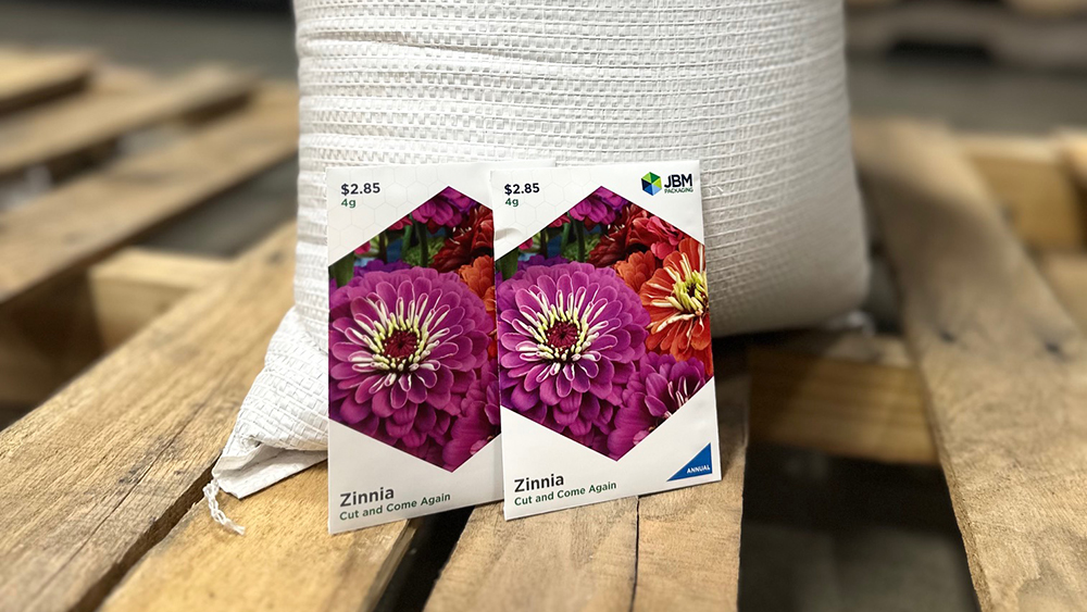 Two JBM branded zinnia seed packets propped against a large sack of seeds sitting on a wooden pallet.