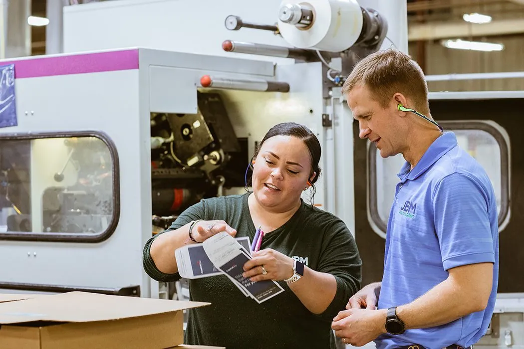 A woman shows a man envelopes in a manufacturing facility.