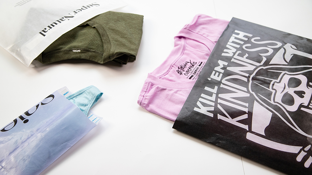 Three glassine bags holding various colorful apparel items. The main glassine bag in view holds a pink T-shirt in a black glassine bag labeled with a skull and the text "Kill 'Em With Kindness."